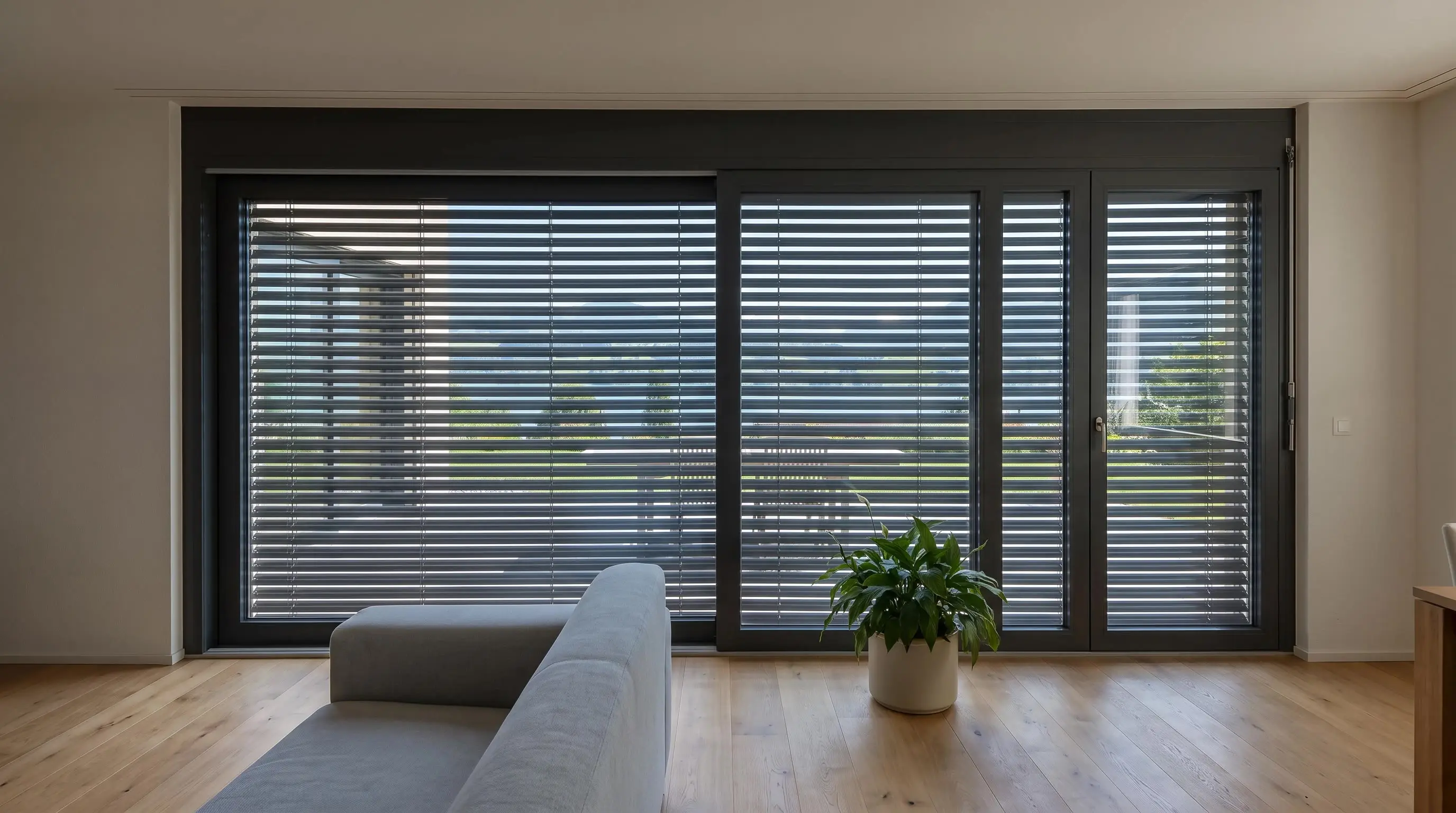 Wohnzimmer mit grosser Fensterfront, aussen sind die anthrazitfarbenen Lamellenstoren ganz unten und geschlossen, der Raum liegt im Schatten