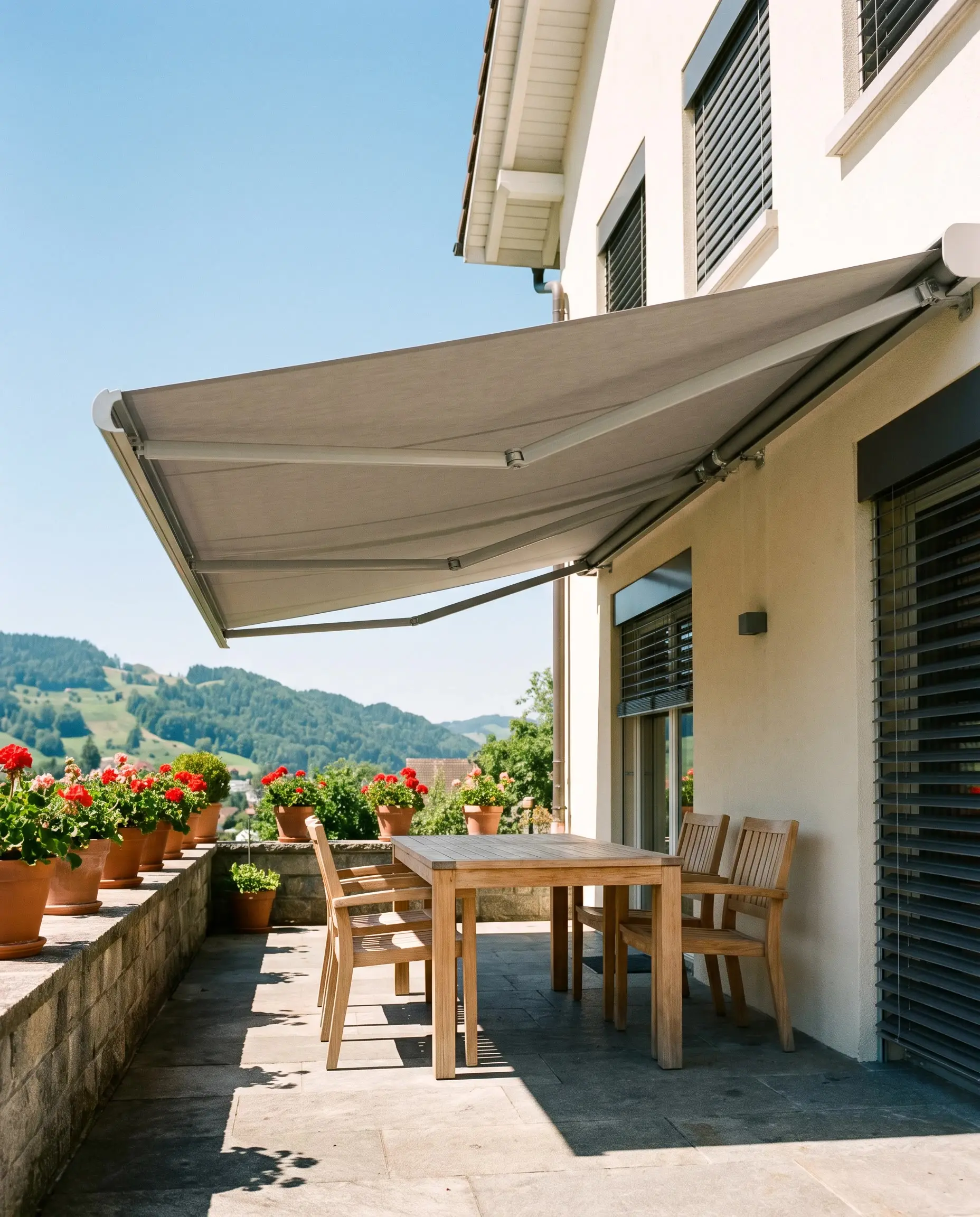 Ausgefahrene graue Gelenkarmmarkise über einer Terrasse mit Holztisch, Stühlen und roten Geranien, dahinter grüne Hügel
