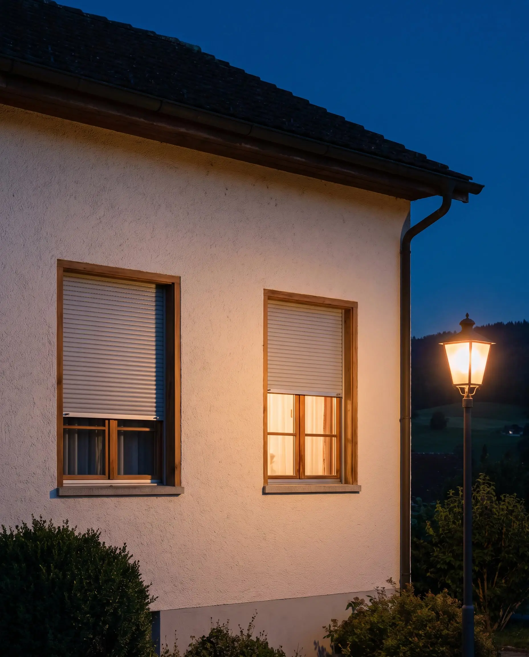 Älteres Haus am Abend: ein Rollladen ganz zu, der andere halb unten mit warmem Licht im Zimmer, daneben eine Strassenlampe